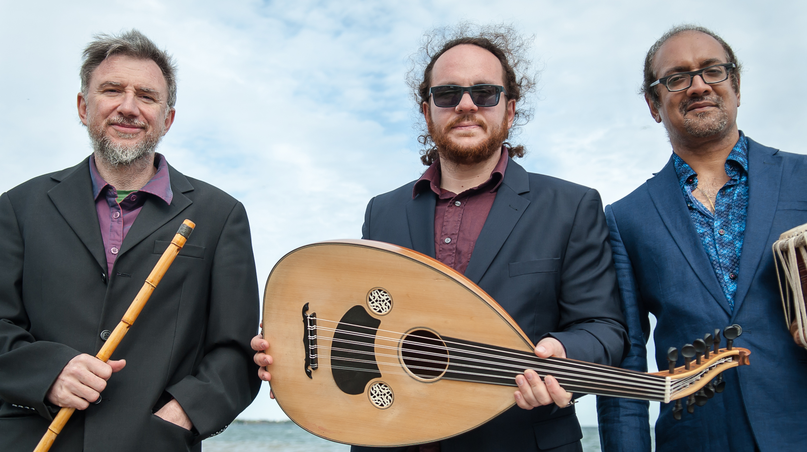 Three men in blazers stand outside holding instruments. A reed-style flute, a guitar-shaped instrumented that is decorated, and a drum.